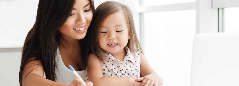 Image of amazing young mom sitting at the table with little cute asian girl at home indoors using laptop computer writing notes to notebook. Looking aside.